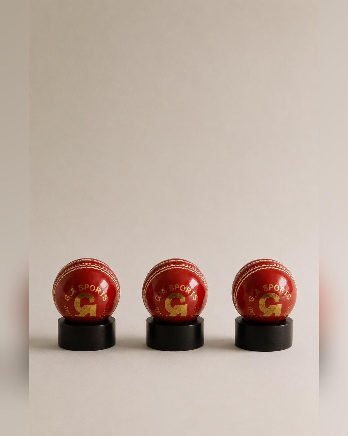 Three red leather cricket balls stamped G.A Sports in gold, each resting on a small black cylindrical stand, seams stitched in white thread.