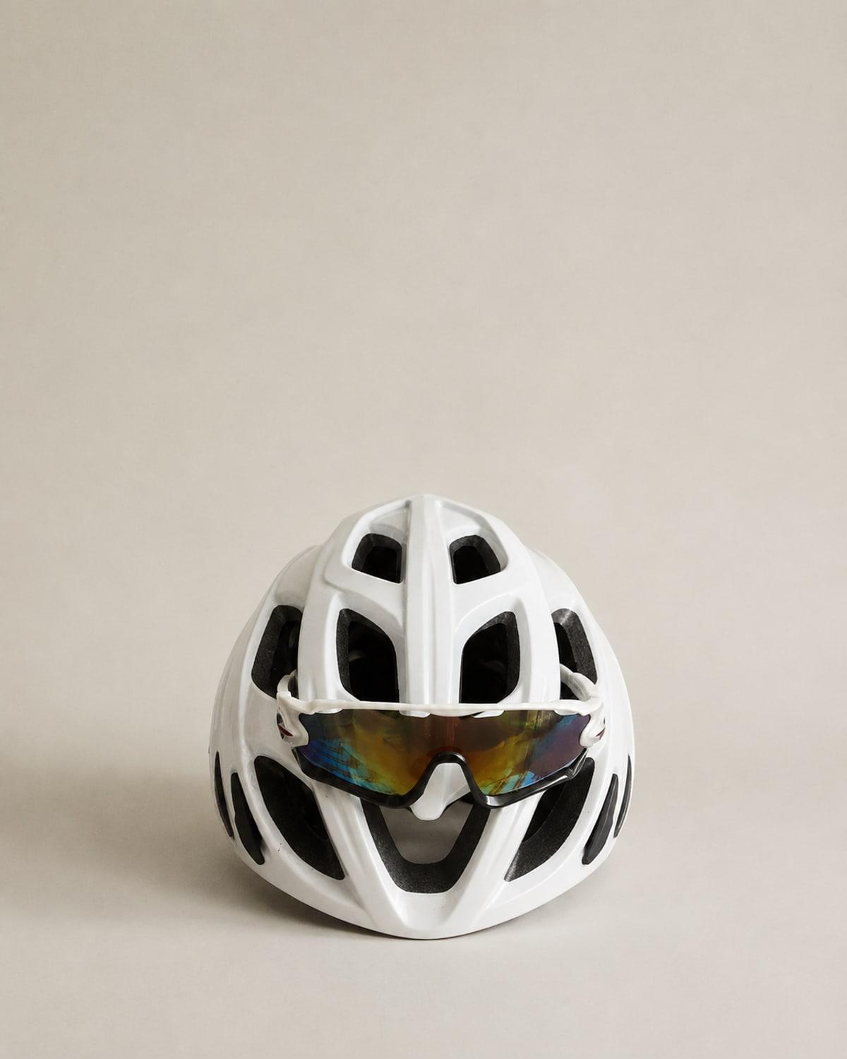 A white vented cycling helmet seen from the front with a pair of wraparound sunglasses hooked into the front vents, their lenses mirrored in rainbow orange and blue.