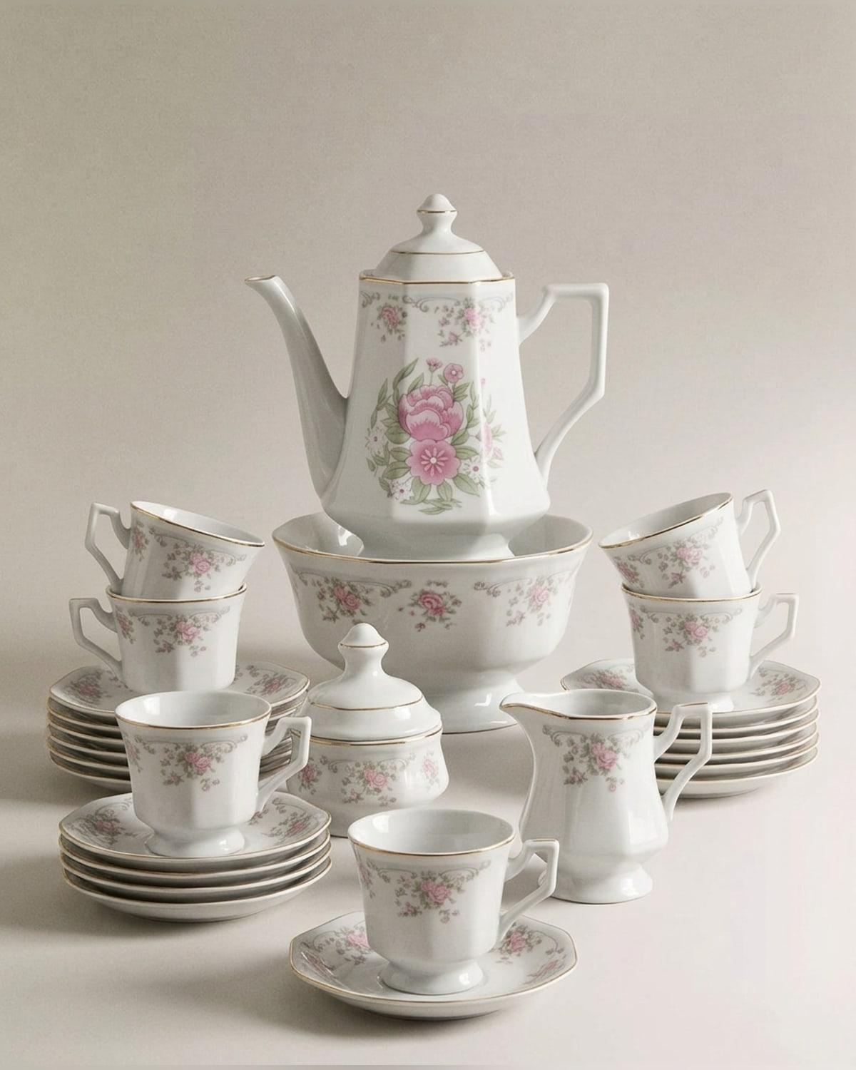 A white porcelain tea service printed with pink roses and gold rims — tall faceted coffee pot, sugar bowl, milk jug, six cups and stacks of saucers and plates arranged around a slop bowl.